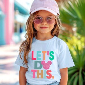 May include: A light blue t-shirt with the text "LET'S DO THIS" in colorful block letters and a glittery pink Mickey Mouse head. The shirt is worn by a child wearing a pink cap and sunglasses.