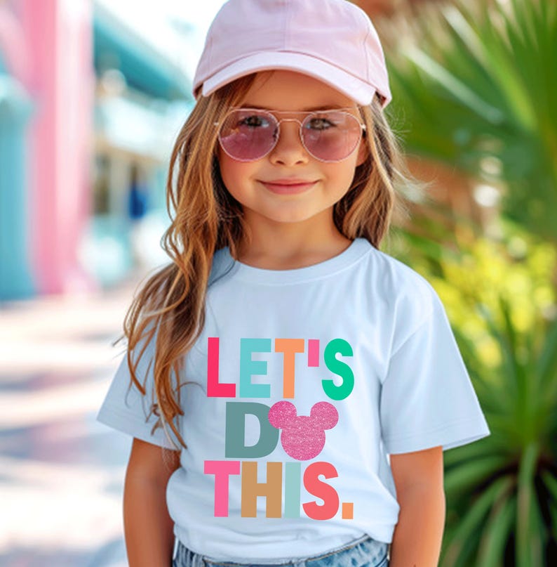 May include: A light blue t-shirt with the text "LET'S DO THIS" in colorful block letters and a glittery pink Mickey Mouse head. The shirt is worn by a child wearing a pink cap and sunglasses.