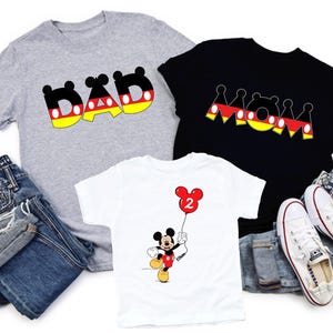 May include: Three t-shirts with graphic designs. A grey t-shirt with the word "DAD" in black, yellow, and red. A black t-shirt with the word "MOM" in black, yellow, and red. A white t-shirt with a Mickey Mouse graphic holding a red balloon.