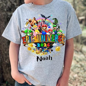 Pu&ograve; includere: Maglietta grigia con una grafica colorata. La grafica include il testo "Epic Universe 2026" e il nome "Noah", insieme a vari personaggi dei cartoni animati e un arcobaleno.