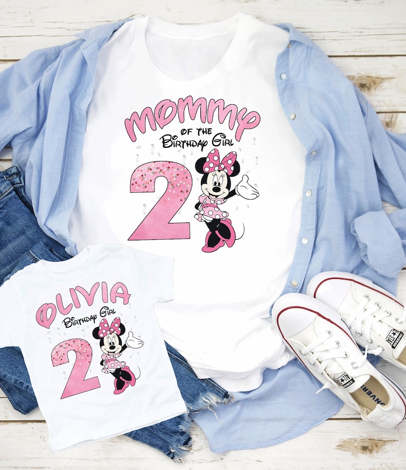 Puede incluir: Camisetas blancas con gr&aacute;ficos de Minnie Mouse. Una camiseta dice "Mam&aacute; de la cumplea&ntilde;era" con un n&uacute;mero 2 rosa y Minnie Mouse. La otra camiseta dice "Olivia Birthday Girl" con un n&uacute;mero 2 rosa y Minnie Mouse. Camisa azul y jeans en el fondo.
