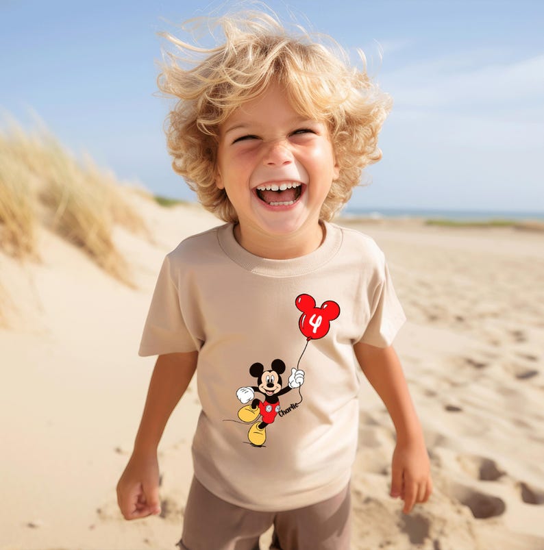 May include: A beige t-shirt with a Mickey Mouse cartoon holding a red balloon with the number 4. The shirt has a personalized name below the character. The child is smiling on a sandy beach.
