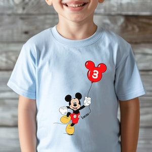 May include: Pale blue t-shirt with a Mickey Mouse design holding a red balloon with the number 3. The cartoon character is wearing red shorts and yellow shoes. The name "Lucas" is written on the shirt.