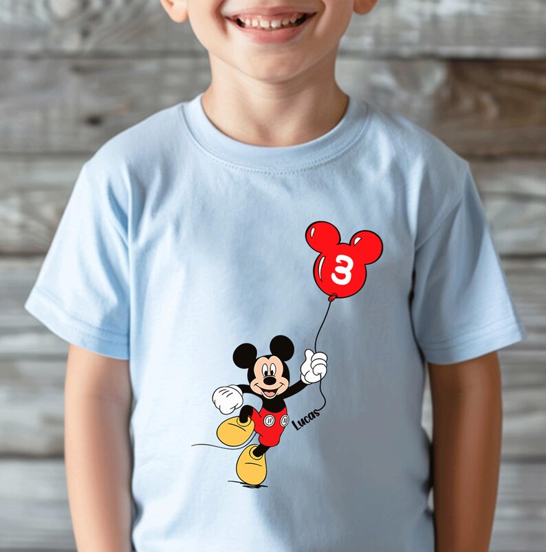 May include: Pale blue t-shirt with a Mickey Mouse design holding a red balloon with the number 3. The cartoon character is wearing red shorts and yellow shoes. The name "Lucas" is written on the shirt.