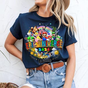 Pu&ograve; includere: T-shirt blu navy con una grafica colorata di personaggi di videogiochi, tra cui Mario e Luigi. Il testo "EPIC UNIVERSE 2026" &egrave; ben visibile. &Egrave; incluso anche un nome personalizzato.