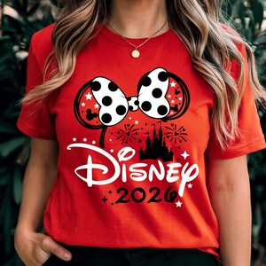 May include: A bright red short-sleeved t-shirt featuring a graphic of a white polka-dot bow with black outlines, a black silhouette of a castle, fireworks, and the white text Disney above the black text 2026, accented with small white stars.