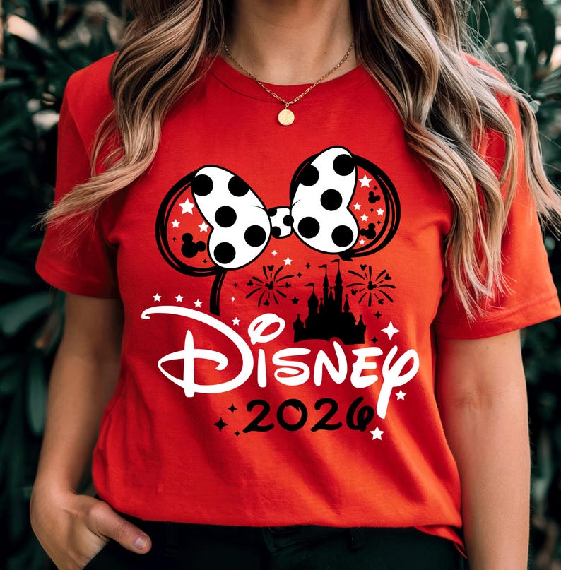 May include: A bright red short-sleeved t-shirt featuring a graphic of a white polka-dot bow with black outlines, a black silhouette of a castle, fireworks, and the white text Disney above the black text 2026, accented with small white stars.