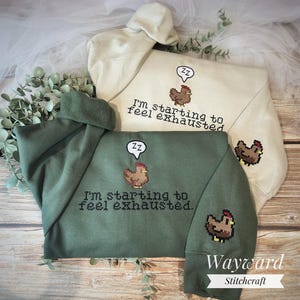 May include: Two hooded sweatshirts, one sage green and one cream, featuring the text "I'm starting to feel exhausted" and a pixelated chicken graphic. The green sweatshirt has a chicken graphic on the sleeve. The brand name "Wayward Stitchcraft" is at the bottom.