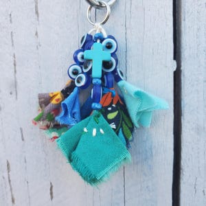 May include: A decorative keychain featuring a central turquoise cross bead, multiple blue and white evil eye beads, and several fabric scraps in shades of teal, blue, and floral patterns with frayed edges, all attached to a silver metal ring.