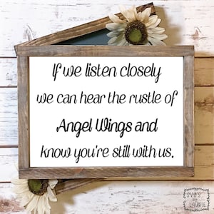 以下が含まれることがあります： 白い木製の看板に黒い文字で「よく耳を傾ければ、天使の羽根の音が聞こえて、あなたはまだ私たちと一緒にいるとわかるでしょう。」と書かれています。