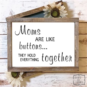 Puede incluir: Un letrero de madera blanco con el texto "Moms are like buttons... They hold everything together". El letrero está enmarcado en un marco de madera rústico y tiene un girasol en la parte superior.