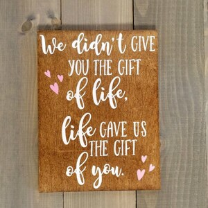 May include: A wooden sign with the text "We didn't give you the gift of life, life gave us the gift of you."  The text is in white with pink hearts around the text.