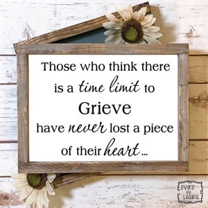 May include: A white wooden sign with black text that reads "Those who think there is a time limit to Grieve have never lost a piece of their heart..." The sign is framed in a rustic wooden frame.