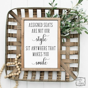 May include: A white sign with black text that reads "Assigned seats are not our style. Sit anywhere that makes you smile." The sign is framed in wood and is sitting in a wooden basket.