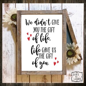 May include: A white wooden sign with black text that reads "We didn't give you the gift of life, life gave us the gift of you." There are small red hearts scattered around the text.