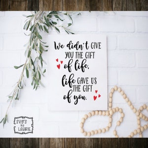 May include: White wooden sign with black text that reads "We didn't give you the gift of life, life gave us the gift of you." There are red hearts around the text.