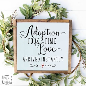 May include: A white sign with a brown wooden frame, featuring the text "Adoption took time Love arrived instantly" in black cursive font with a small red heart below.