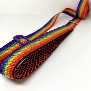 May include: A rainbow-striped adjustable strap featuring a vibrant pattern of red, orange, yellow, green, blue, and purple. The strap includes a purple plastic slider and a black mesh lining on the underside, designed for carrying or securing items.