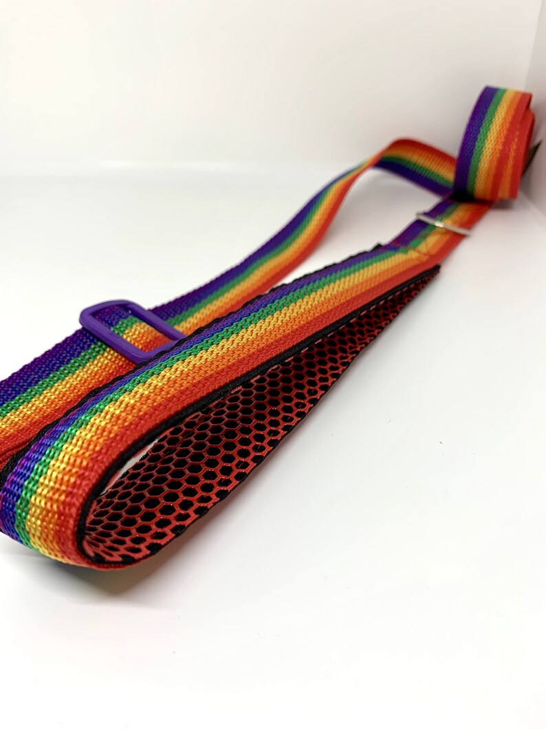 May include: A rainbow-striped adjustable strap featuring a vibrant pattern of red, orange, yellow, green, blue, and purple. The strap includes a purple plastic slider and a black mesh lining on the underside, designed for carrying or securing items.