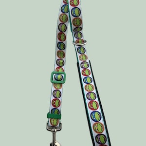 May include: White dog lead with a repeating pattern of colourful turtle faces. The lead has a green buckle and a silver metal clasp. The edges of the lead are trimmed in black.