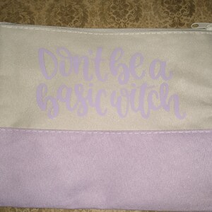 May include: A rectangular cosmetic bag with a light gray upper section and a lavender lower section. The text "Don't be a basic witch" is printed in lavender script across the upper section. The bag has a silver zipper.