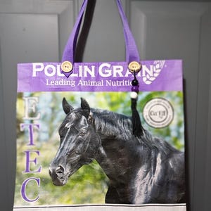 May include: A tote bag made from a feed bag, featuring a black horse graphic. The bag has purple handles and accents, with the text "POLLIN GRAN" and "SENIOR LOW" visible. Decorative buttons and a tassel are attached.