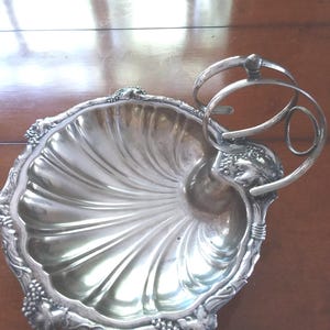 May include: A silver-colored, nautilus-shaped serving dish with a decorative handle. The dish has a scalloped edge with grape cluster accents. The handle is composed of two curved rings and a small ball. The dish is likely made of metal.