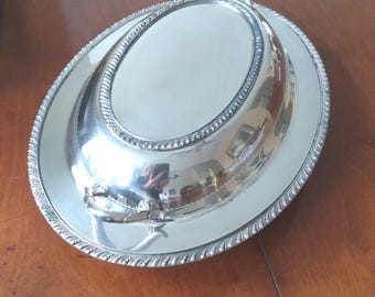 May include: A silver-colored serving dish with a lid and a handle. The dish has an oval shape with a decorative beaded edge and a rope-like border. The lid has a handle for easy lifting. The dish is sitting on a wooden surface.