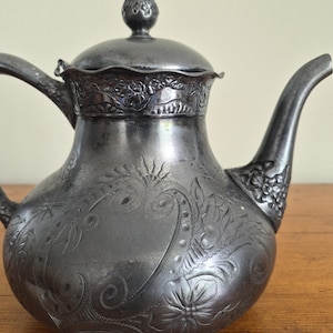 May include: Antique silver-colored teapot with intricate floral and swirl engravings. The teapot features a curved spout, a handle, and a lid with a decorative knob. The design includes detailed patterns across the body and lid, showcasing a vintage aesthetic.