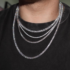 May include: A layered set of four silver-toned tennis necklaces featuring round, clear, faceted stones set in a continuous row. The necklaces vary in length and stone size, creating a cascading effect against a dark grey crew neck t-shirt.