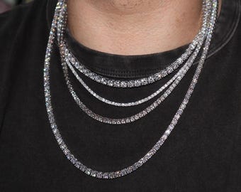 May include: A layered set of four silver-toned tennis necklaces featuring round, clear, faceted stones set in a continuous row. The necklaces vary in length and stone size, creating a cascading effect against a dark grey crew neck t-shirt.