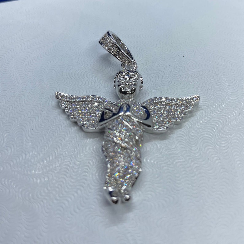 May include: A silver-colored angel pendant with outstretched wings and hands clasped in prayer. The pendant is encrusted with numerous small, sparkling stones, and has a loop at the top for a chain. The angel's wings are spread wide, and the details are finely crafted.