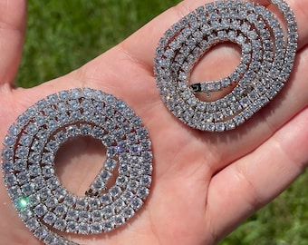 May include: Two silver tennis chains with clear cubic zirconia stones. The chains are laid out on a hand, showing the detail of the stones.