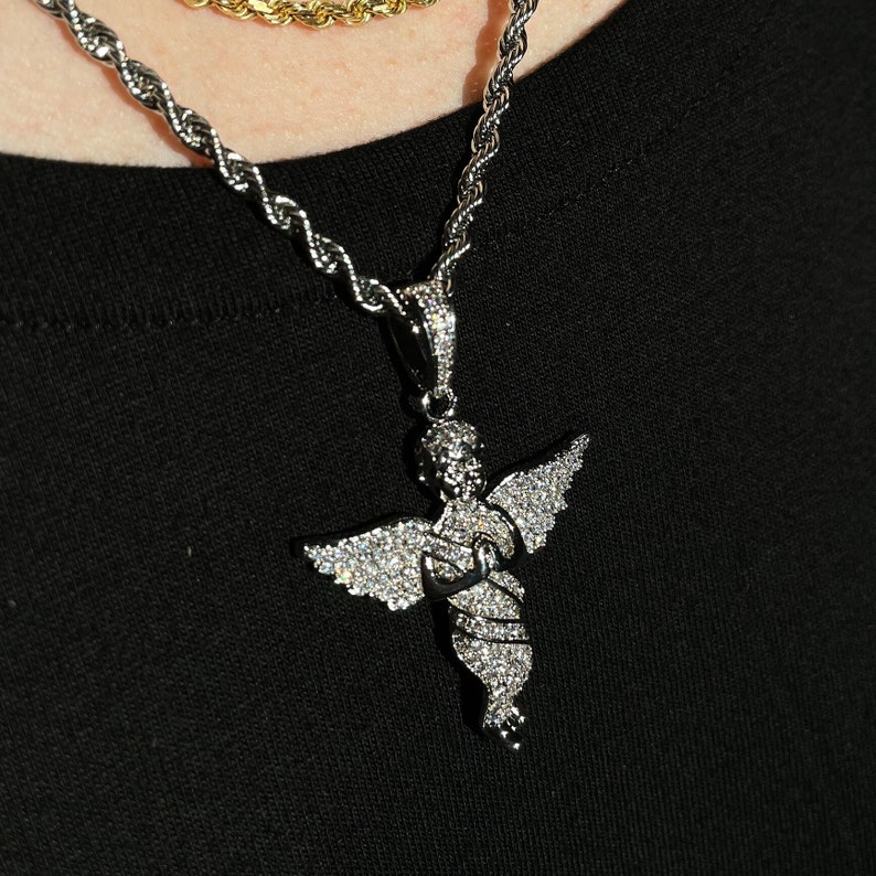 May include: Silver-toned angel pendant necklace with a rope chain. The angel pendant is encrusted with small, sparkling stones. A gold-toned rope chain is visible in the background. The necklace is worn against a black background.