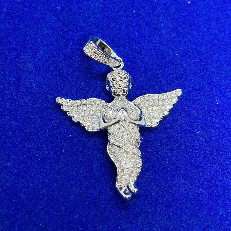 May include: A silver angel pendant with a halo and outstretched wings, encrusted with small, sparkling stones. The angel is depicted with hands clasped in prayer. The pendant hangs from a silver bail, set against a blue background.