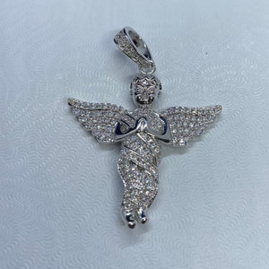 May include: A silver-colored angel pendant with a halo and outstretched wings. The angel is adorned with numerous small, sparkling stones, giving it a brilliant appearance. The pendant is designed with intricate details and is suspended from a matching bail.