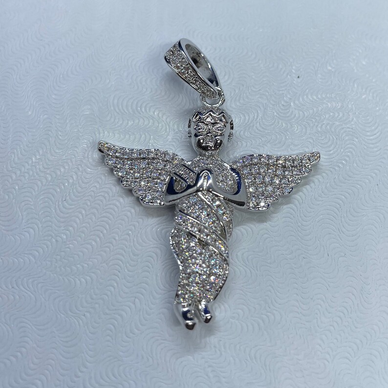 May include: A silver-colored angel pendant with a halo and outstretched wings. The angel is adorned with numerous small, sparkling stones, giving it a brilliant appearance. The pendant is designed with intricate details and is suspended from a matching bail.