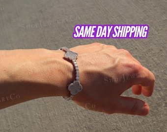 May include: A silver-toned bracelet featuring a series of small, round, clear stones set in a chain, interspersed with two prominent four-leaf clover-shaped charms covered in pavé-set clear stones. The bracelet is worn on a wrist.
