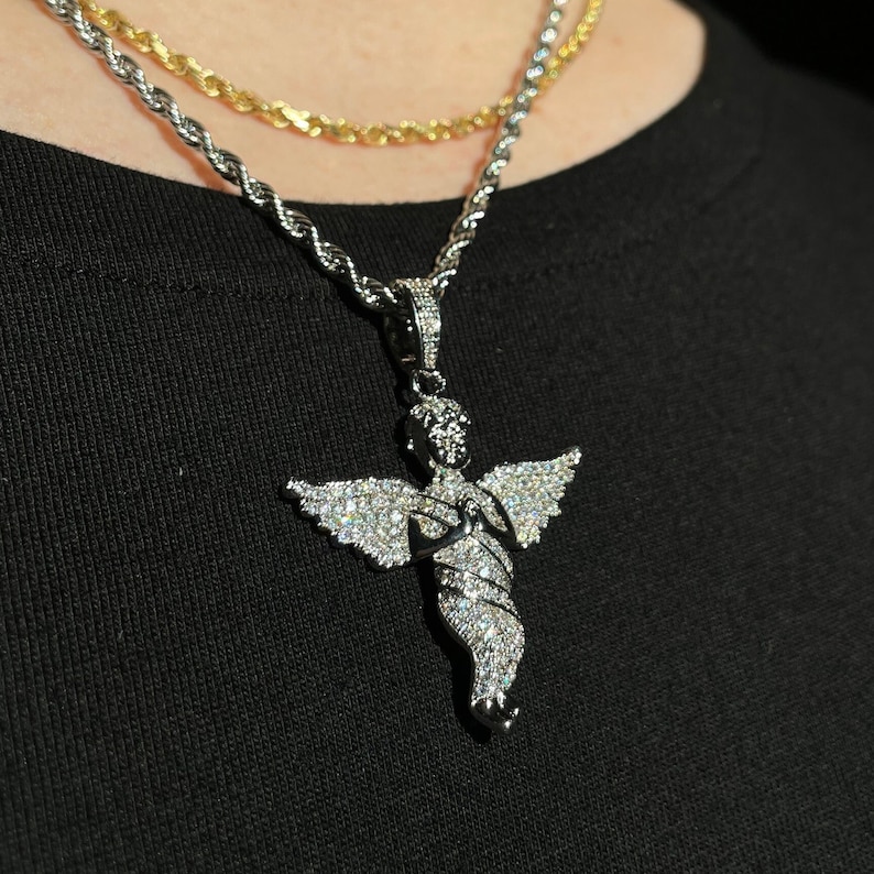 May include: A silver-toned angel pendant necklace with sparkling clear stones. The angel has outstretched wings and is suspended from a silver rope chain. A gold rope chain is also visible in the background. The necklace is worn on a black shirt.