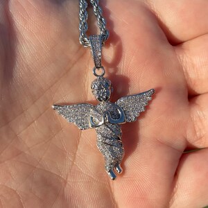 May include: A silver-colored angel pendant necklace with a rope chain. The angel pendant is encrusted with small, clear stones, and has outstretched wings and clasped hands. The necklace is displayed against a neutral background.