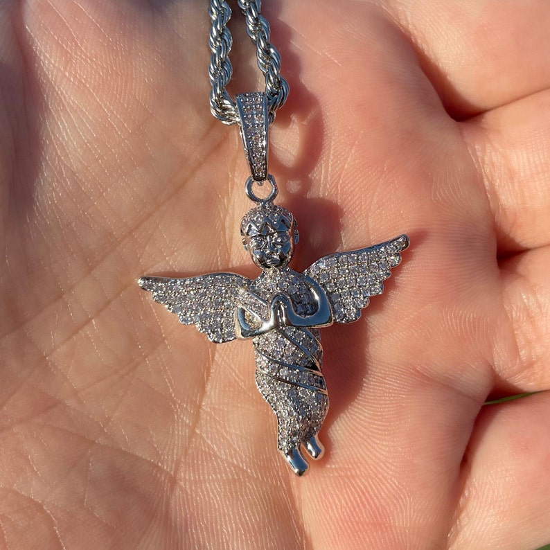 May include: A silver-colored angel pendant necklace with a rope chain. The angel pendant is encrusted with small, clear stones, and has outstretched wings and clasped hands. The necklace is displayed against a neutral background.