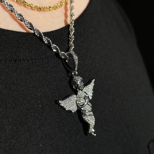 May include: Silver-toned angel pendant necklace with a diamond-like encrusted design. The angel pendant is suspended from a silver rope chain. A gold rope chain is visible in the background. The necklace is worn on a black shirt.