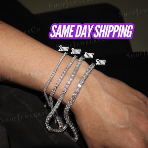 May include: Four silver-toned tennis bracelets with clear stones, labeled with widths of 2mm, 3mm, 4mm, and 5mm. The bracelets are arranged in a row on a wrist, showcasing the varying thickness of the metal chains and the size of the stones.