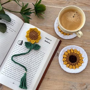May include: A crochet sunflower bookmark with a green stem and tassel, placed on an open book titled Chapter 10 Believe, alongside two round crochet sunflower coasters, one of which supports a light yellow ceramic mug filled with coffee.