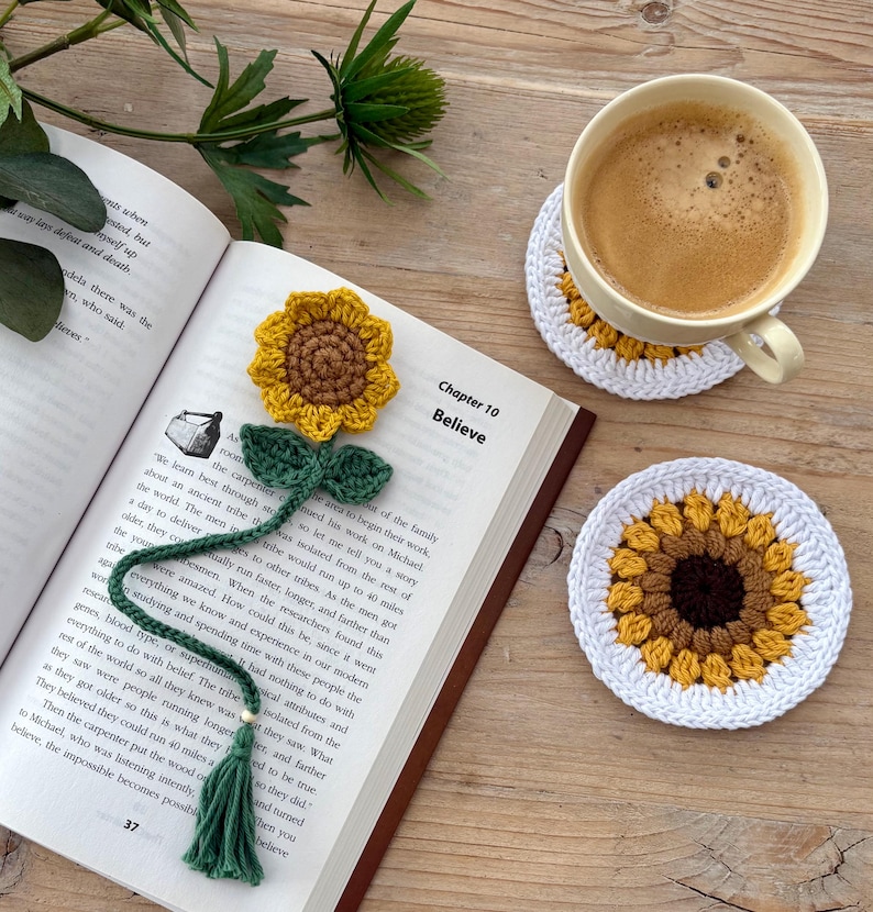 May include: A crochet sunflower bookmark with a green stem and tassel, placed on an open book titled Chapter 10 Believe, alongside two round crochet sunflower coasters, one of which supports a light yellow ceramic mug filled with coffee.
