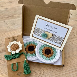 May include: A brown cardboard gift box containing two round crochet coasters with brown, light green, and white sunflower patterns. A matching crochet sunflower bookmark is attached to a card, and a thank you note features a small heart and pompom.