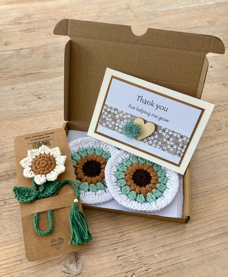 May include: A brown cardboard gift box containing two round crochet coasters with brown, light green, and white sunflower patterns. A matching crochet sunflower bookmark is attached to a card, and a thank you note features a small heart and pompom.