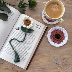 May include: A crochet sunflower bookmark with a green stem and tassel resting on an open book, alongside two round crochet coasters in white, pink, and brown. The book page shows the text Chapter 10 Believe and printed paragraphs of text.