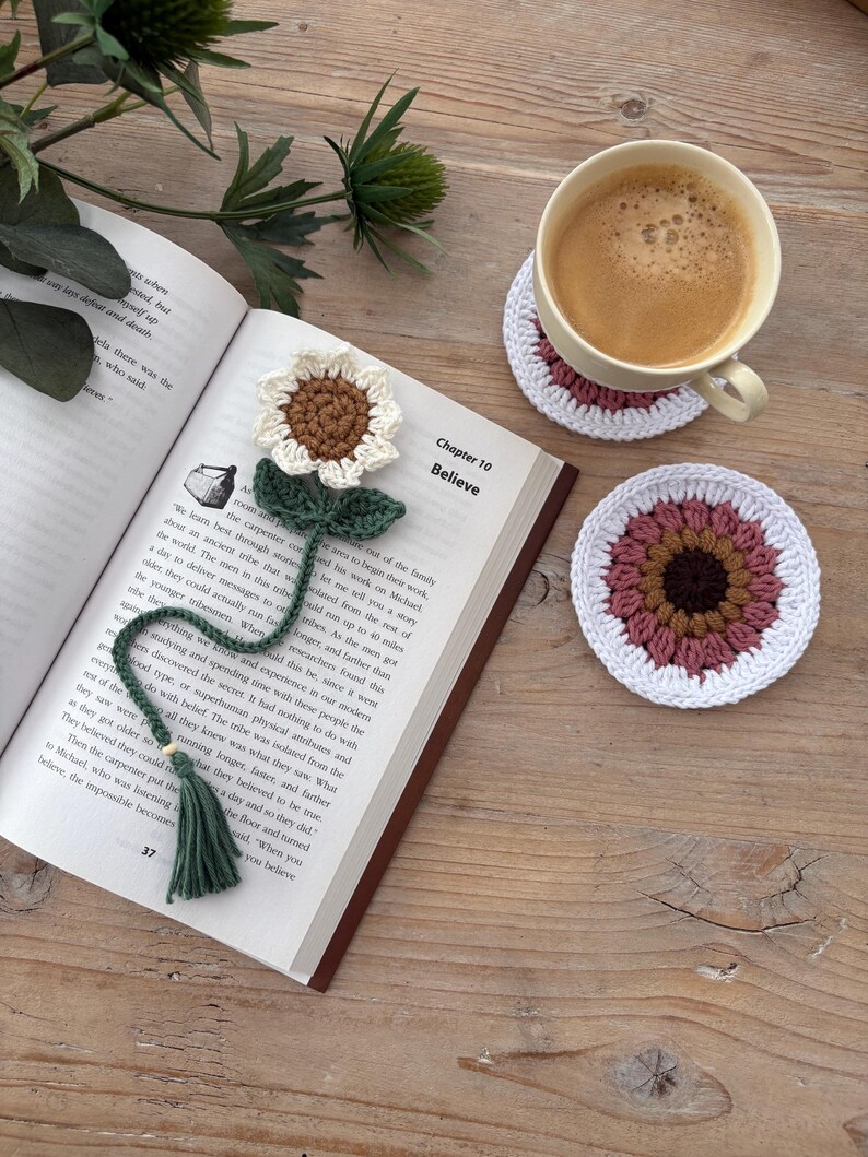 May include: A crochet sunflower bookmark with a green stem and tassel resting on an open book, alongside two round crochet coasters in white, pink, and brown. The book page shows the text Chapter 10 Believe and printed paragraphs of text.