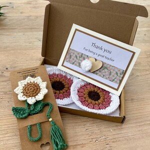 May include: A brown cardboard gift box containing two round crocheted coasters with pink and brown centres and white edges, and a card that reads Thank you For being a great teacher. A crocheted sunflower bookmark is attached to a brown card.